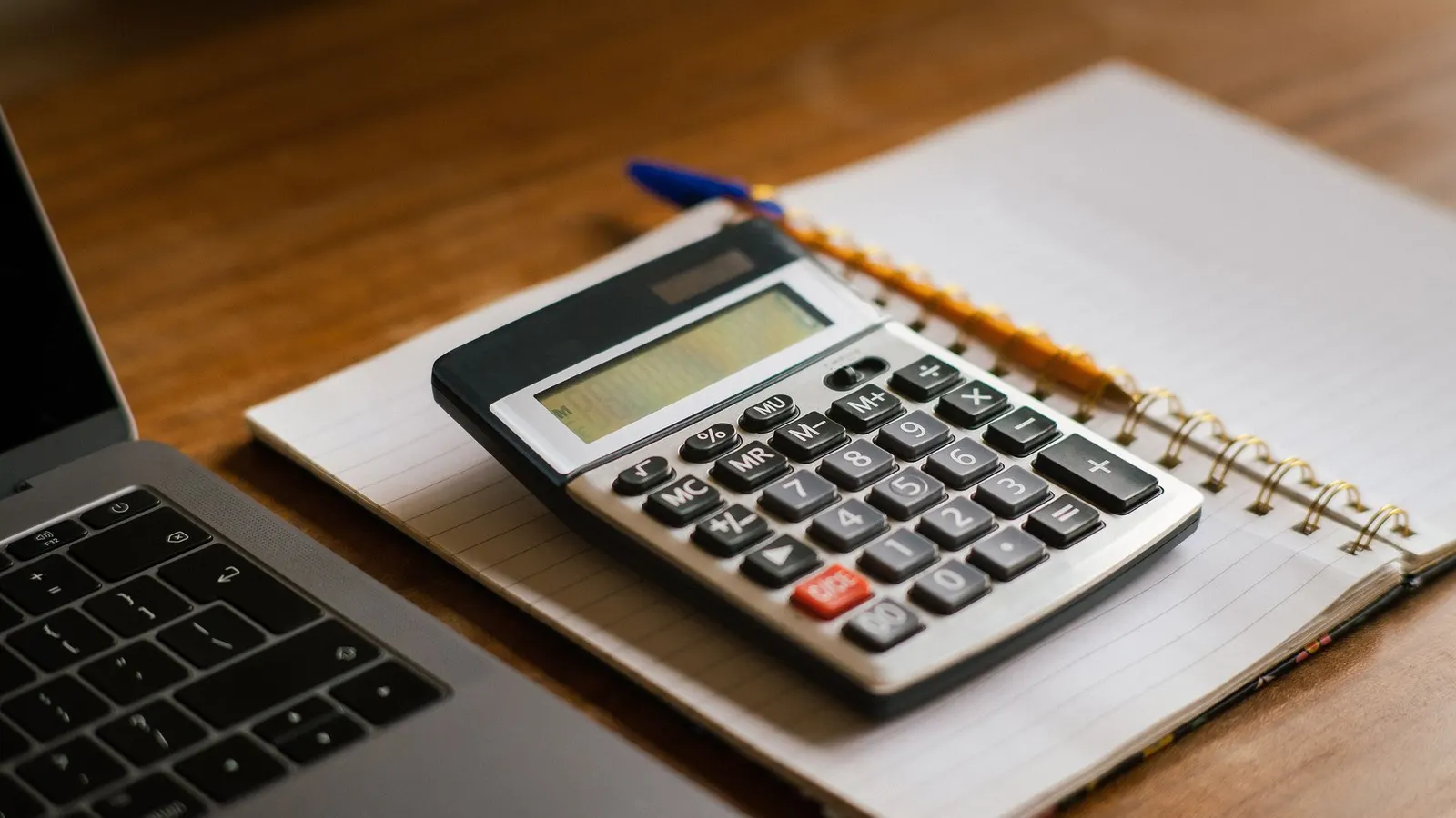 Una calculadora sobre un cuaderno, junto a una notebook en un escritorio de madera