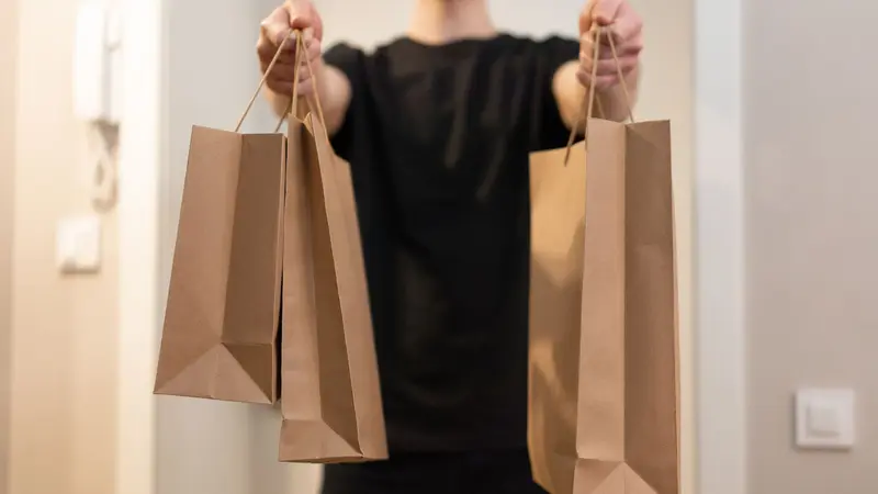 Una persona de remera negra extiende los brazos para entregar tres bolsas de papel madera con un pedido