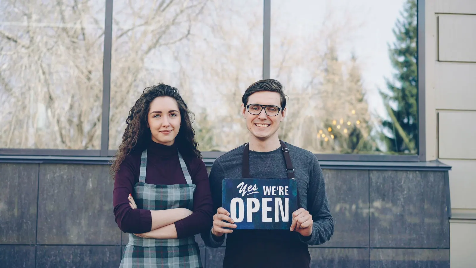 Una pareja con delantales frente a su local; él sostiene un cartel que dice que están abiertos