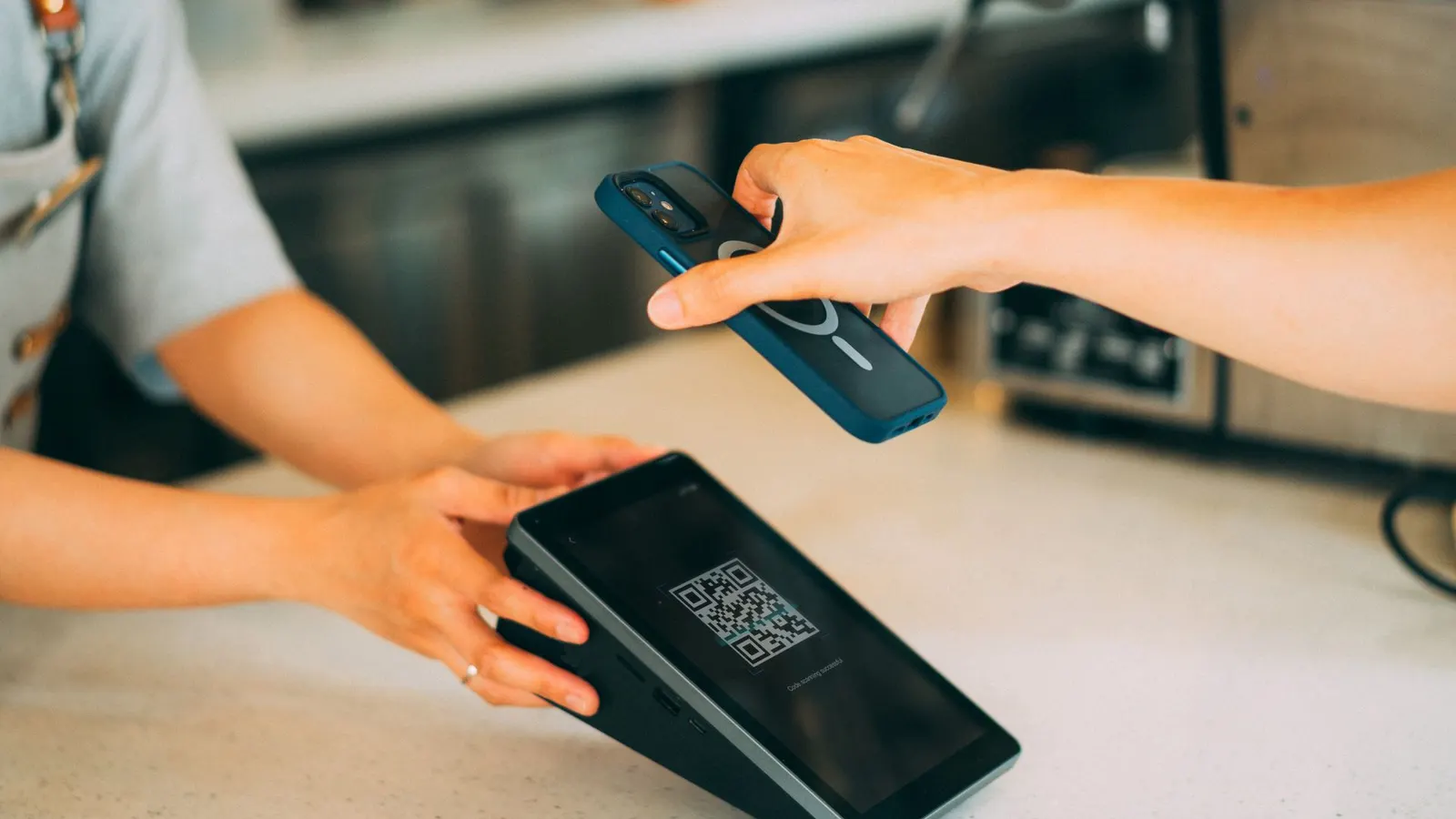 Un celular se acerca a una terminal con un código QR en la pantalla, sobre el mostrador de un comercio
