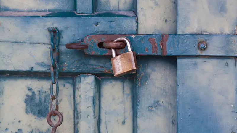 Puerta de madera azul despintada, trabada con un candado y una cadena oxidada
