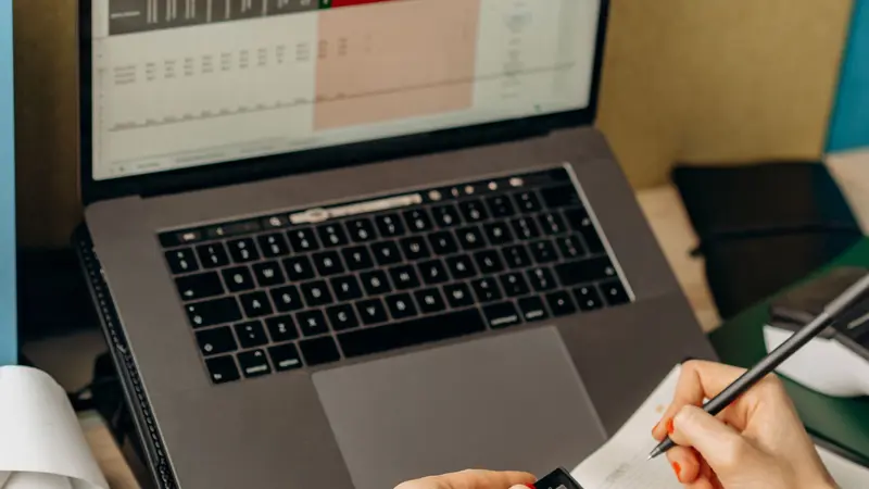 Manos escribiendo notas junto a una laptop con datos en pantalla