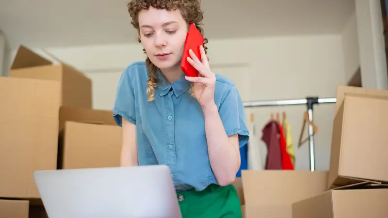 Una emprendedora habla por celular frente a su computadora portátil, entre cajas de envío y un perchero con ropa